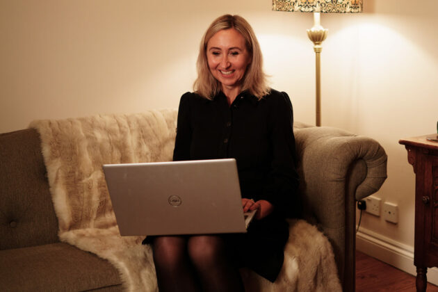A woman sat on a sofa in a cosy environment with a laptop.