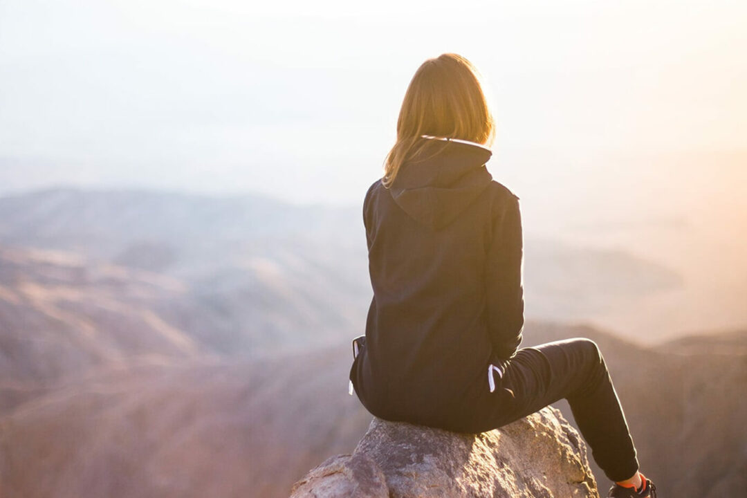 A woman in wearing hiking clothes sat at the top of a mountain looking out onto a beautiful view.