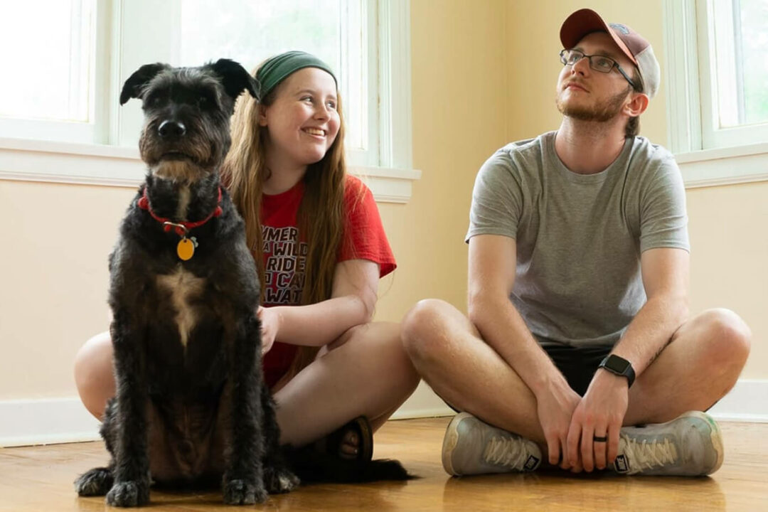 A young couple with their dog sat in an empty room smiling.