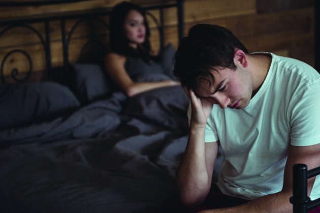 man sitting on bedside looking unhappy with woman in bed in background