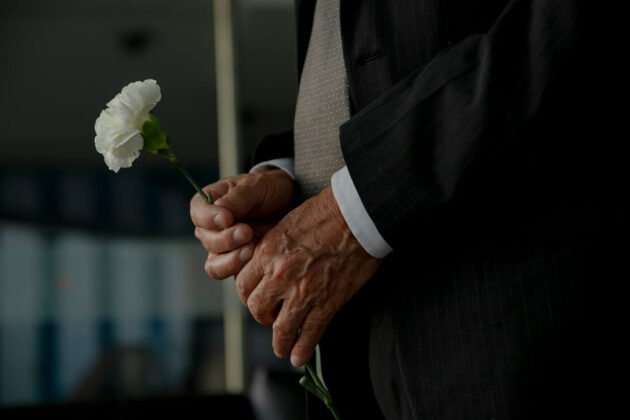 A close up of an elderly mans hands holding a white flower wearing a black suit.