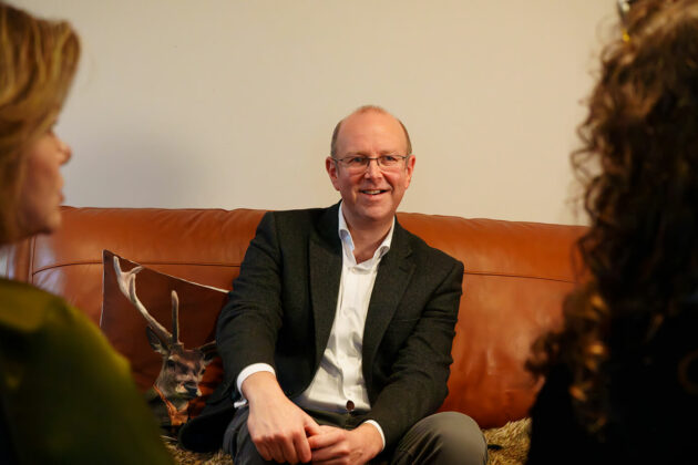 A man sat on a leather sofa - the shot is framed from over the shoulder of two women he is speaking to.